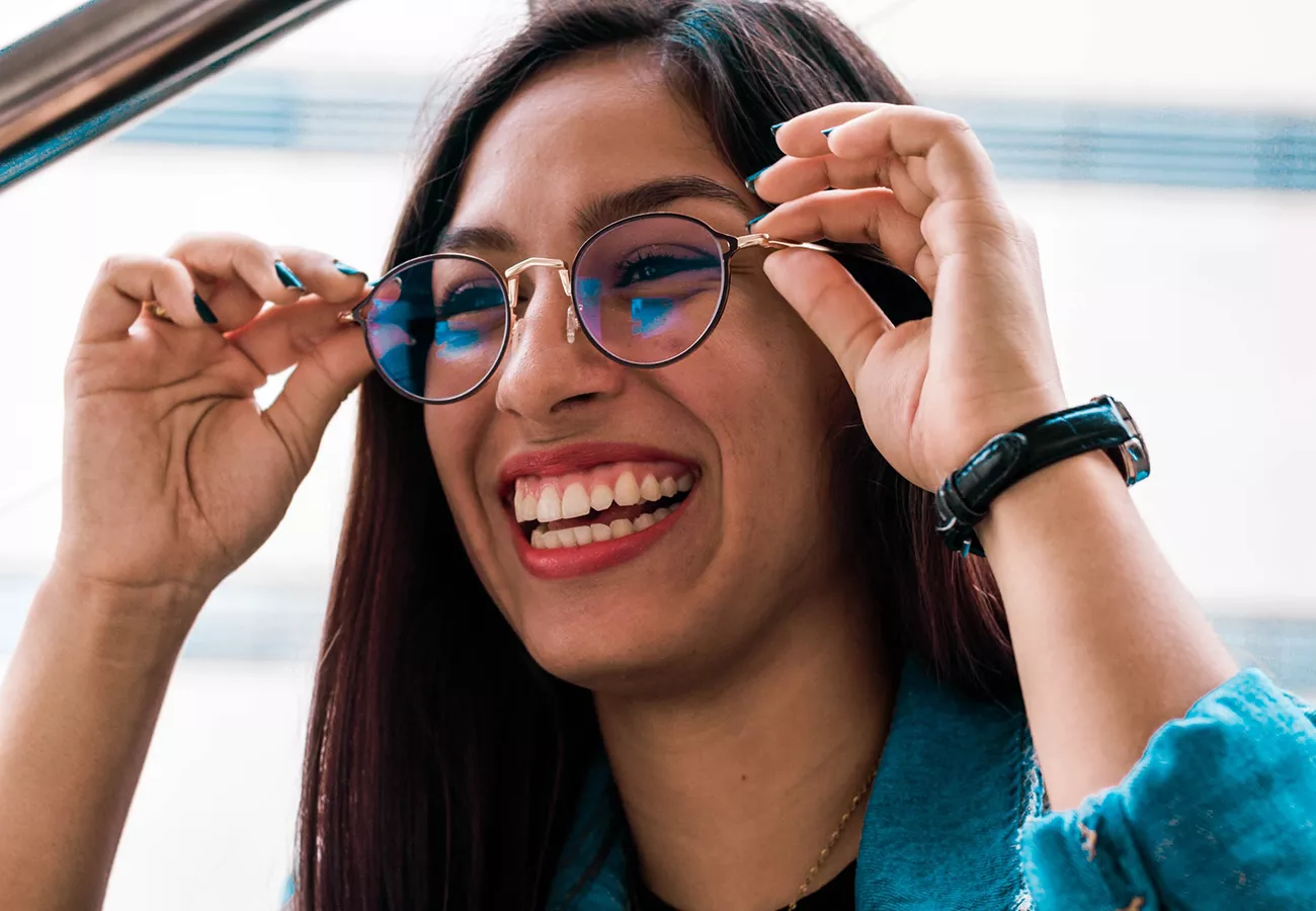 A woman with long dark hair adjusts her sunglasses while outdoors. She is wearing a blue jacket and a black wristwatch. The background features a blurred urban setting with horizontal lines.